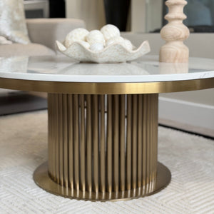 A round coffee table with a white marble top and a fluted gold metal pedestal base, styled in a modern living room setting with a bowl of white decorative spheres and a wooden candlestick holder.