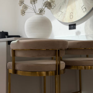 Beige chair with gold legs in front of a round table with a vase and clock in a room.