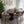 A modern dining set featuring a round wooden table and six upholstered chairs with curved backs, shown in a styled room setting with a patterned rug, framed wall art, and a potted plant.