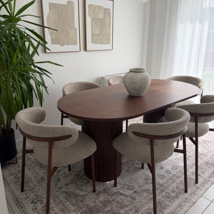 A modern dining set featuring a round wooden table and six upholstered chairs with curved backs, shown in a styled room setting with a patterned rug, framed wall art, and a potted plant.