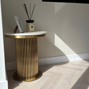 A modern round side table with a fluted gold metal base and a white marble top, styled in a minimalist room with light wood herringbone flooring and a wall-mounted TV.