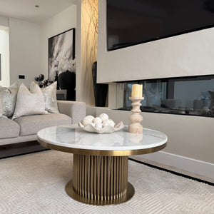 A modern round coffee table with a white marble top and a fluted gold pedestal base, styled in a contemporary living room next to a light gray sofa and a fireplace.