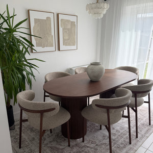 A modern dining set featuring a round wooden table and six upholstered chairs with curved backs, shown in a bright dining room setting with a patterned rug, wall art, and a plant.