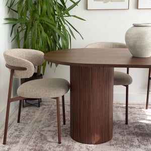 A modern dining room setup featuring a round wooden table with a fluted pedestal base and two upholstered chairs with curved backs, styled with a large potted plant and a ceramic vase.