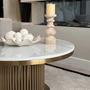 A round coffee table with a white marble top and fluted gold base, styled with a wooden candlestick holder and a decorative bowl of white decorative balls in a modern living room setting.