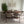 A dining area scene showing a round wooden dining table with multiple upholstered chairs in a neutral beige and brown color palette, set on a patterned gray rug against a white wall with framed abstract art and a hanging white chandelier.