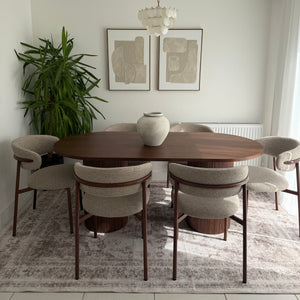 A dining area scene showing a round wooden dining table with multiple upholstered chairs in a neutral beige and brown color palette, set on a patterned gray rug against a white wall with framed abstract art and a hanging white chandelier.