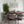 A modern dining room setup featuring an oval wooden dining table surrounded by six upholstered chairs with dark wood frames, set on a distressed gray patterned rug. A large green potted plant stands on the left, and two framed abstract prints hang on the white wall above.