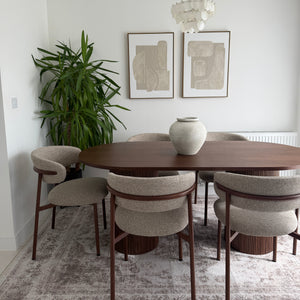 A modern dining room setup featuring an oval wooden dining table surrounded by six upholstered chairs with dark wood frames, set on a distressed gray patterned rug. A large green potted plant stands on the left, and two framed abstract prints hang on the white wall above.