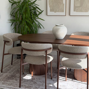 A modern dining room scene featuring a dark wood oval table surrounded by six upholstered dining chairs with curved backs and wooden frames. A large green potted plant stands in the corner, and two framed abstract prints hang on the wall above the table.