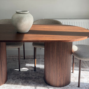 A modern dining table with a warm wood top, a dark wood center stripe, and fluted cylindrical pedestal legs, shown in a styled dining room setting with chairs and a ceramic vase.