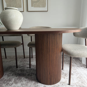 A modern dining table with a fluted dark brown wood grain surface and matching dark brown legs, surrounded by upholstered dining chairs in a neutral beige fabric, set against a textured gray and white patterned wallpaper.