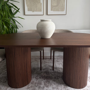 A modern dining table with two ribbed cylindrical dark brown pedestal legs, styled in a minimalist dining room setting with two framed abstract prints on the wall, a white vase, and a green plant.