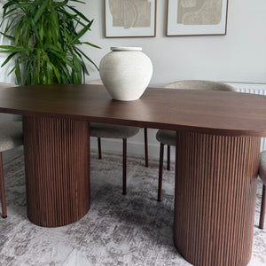 A modern dining room scene showing a dark wood table and matching ribbed bar stools, with a white vase on the tabletop, a green potted plant, and two framed abstract prints on a light wall.