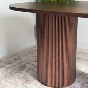 A modern oval wooden dining table with a fluted cylindrical pedestal base, shown in a styled interior setting with a green plant and patterned rug.