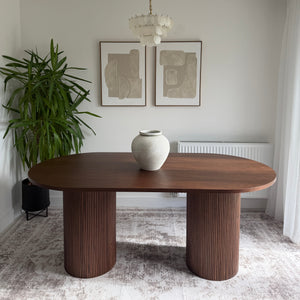 A modern round wooden dining table with two thick ribbed cylindrical legs, shown in a styled interior setting with a white vase on top, a green plant, framed wall art, and a textured wall.