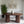 A modern round wooden coffee table with two thick ribbed cylindrical legs and a white ceramic vase on top, shown in a styled interior setting with framed art, a plant, and a textured wall.