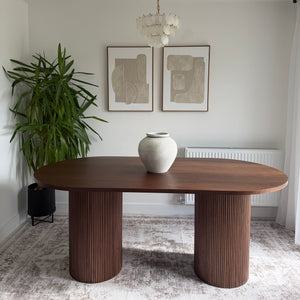 A modern round wooden coffee table with two thick ribbed cylindrical legs and a white ceramic vase on top, shown in a styled interior setting with framed art, a plant, and a textured wall.
