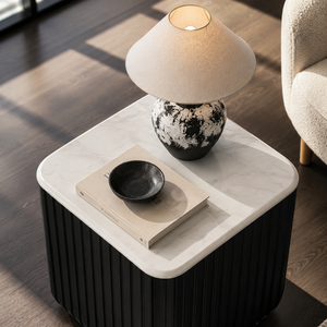 A modern square side table with a black ribbed base and a white marble top, styled in a living room setting with a lit table lamp, a book, and a small bowl on top.